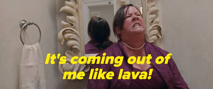 Woman in formal attire, grimacing in discomfort, stands in a bathroom against an ornate mirror