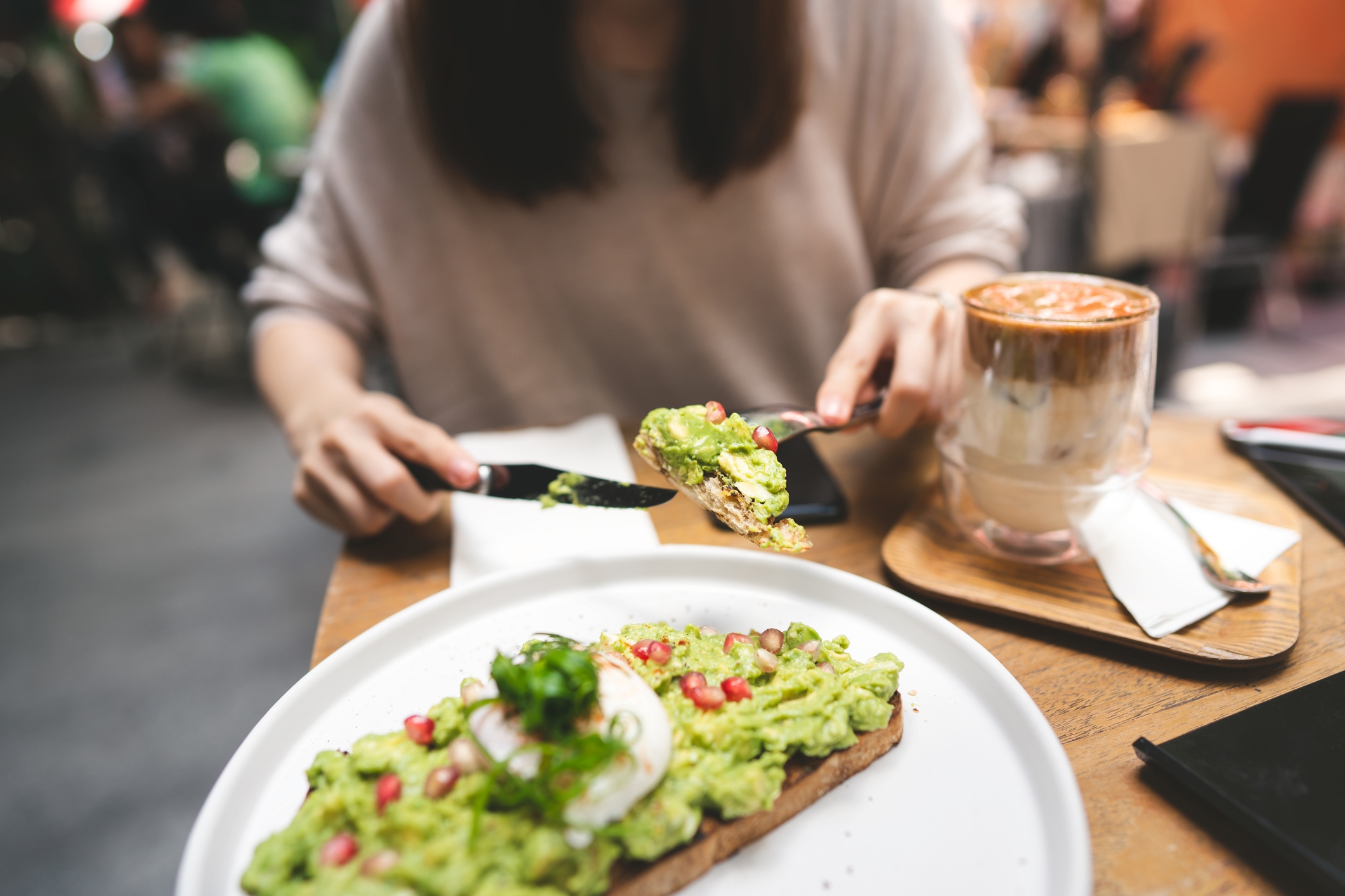 Person eating avocado toast with a poached egg and pomegranate arils, accompanied by a latte, at a café table