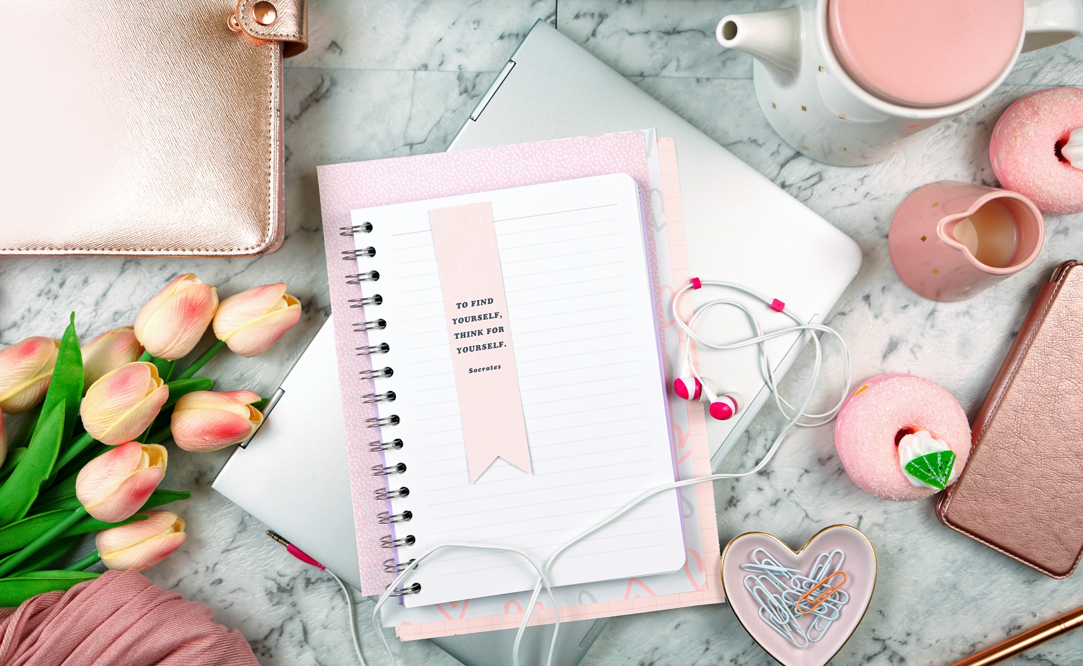 Open notebook with a ribbon bookmark on marble surface, surrounded by tulips, a teapot, earphones, and pink items