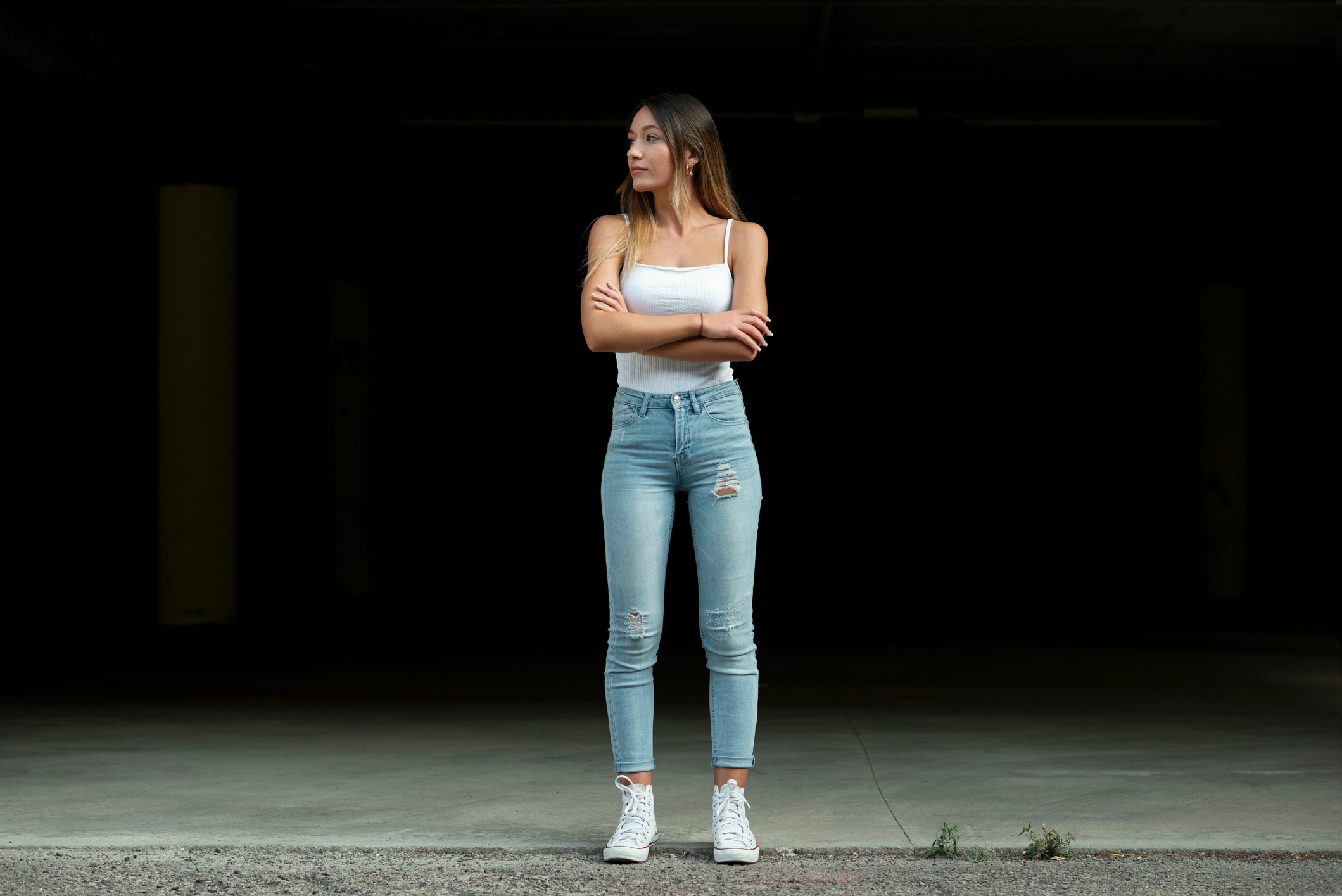 Person stands with crossed arms, wearing a sleeveless top, fitted jeans, and sneakers, looking to the side against a dark background
