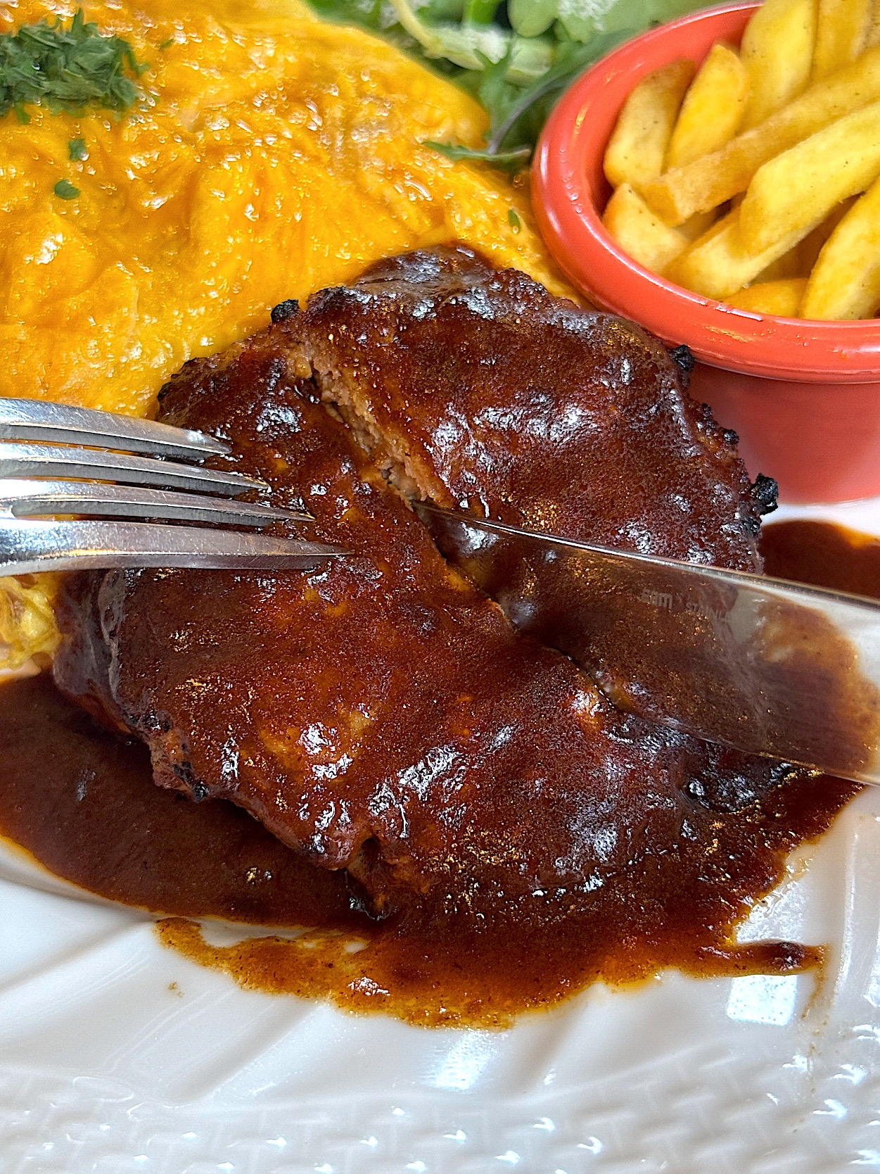 A plate with hamburger steak topped with brown sauce, a fork cutting it, accompanied by fries in a red dish and an omelette