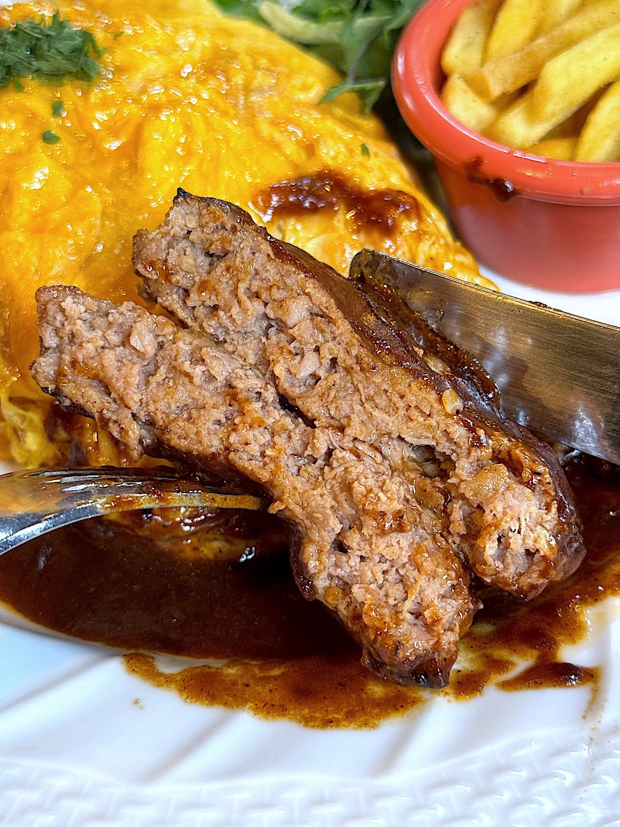 A juicy hamburger steak sliced in half, served with fries and omelet on a plate, drizzled with sauce