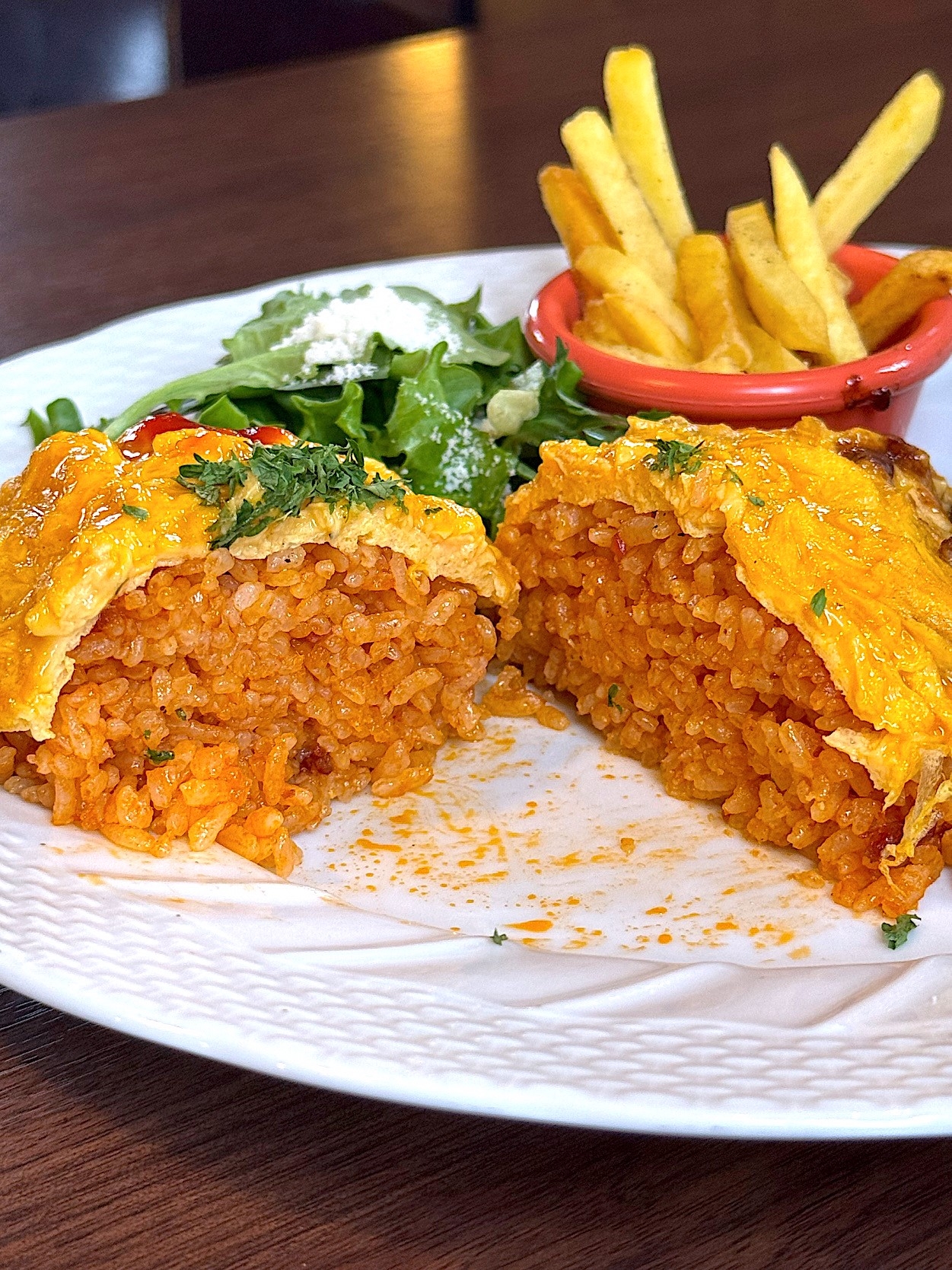 Close-up of an omelet rice dish cut open, revealing seasoned rice inside. Served with fries and a side salad on a white plate