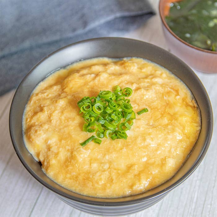 クリーミーな卵料理の上に刻みネギがのった丼。背景にスープの入ったカップ。
