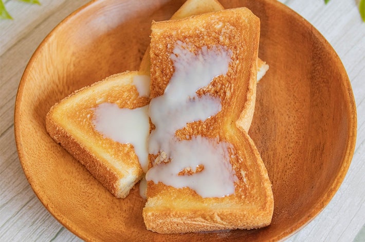 木の皿にトースト2枚が載り、上に練乳がかかっています。バックには緑の葉が見えます。