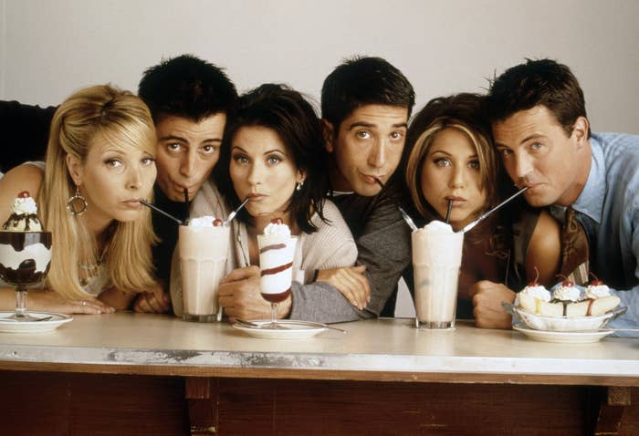 The cast of Friends sitting at a counter, each sipping milkshakes, looking at the camera playfully