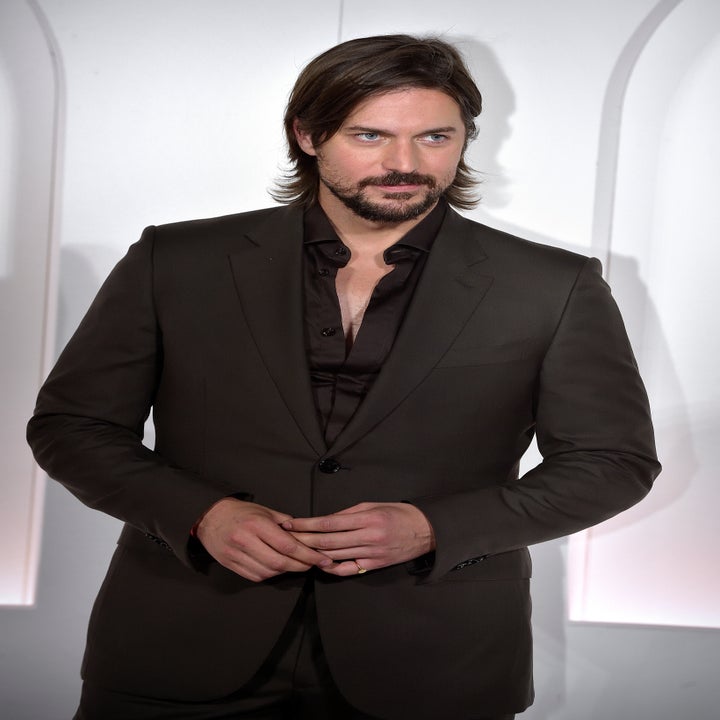 Man in dark suit with open-collar shirt, standing against a white backdrop, gazing thoughtfully ahead