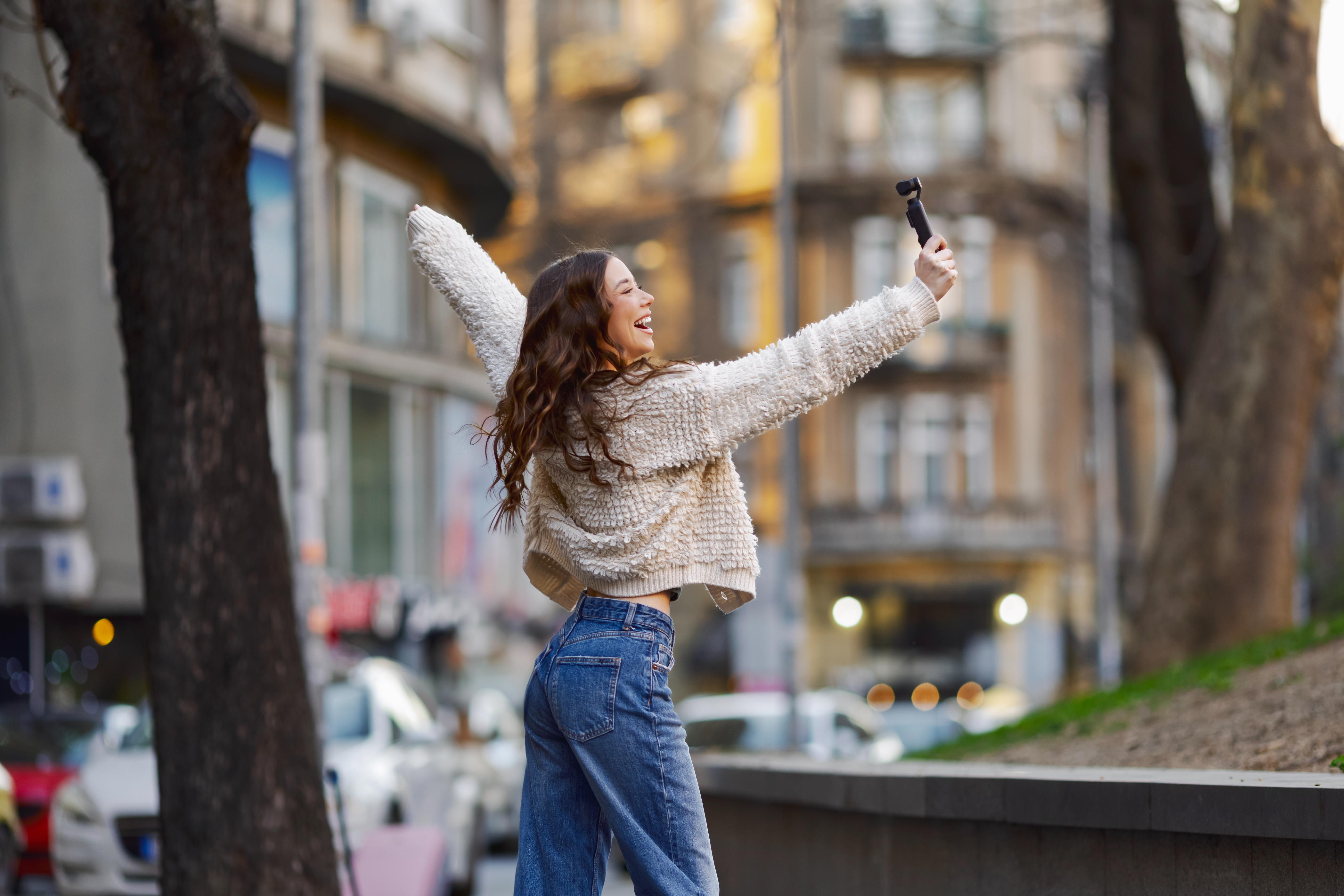 Person outdoors, smiling and holding a phone for a selfie, wearing a textured jacket and jeans, with city buildings in the background