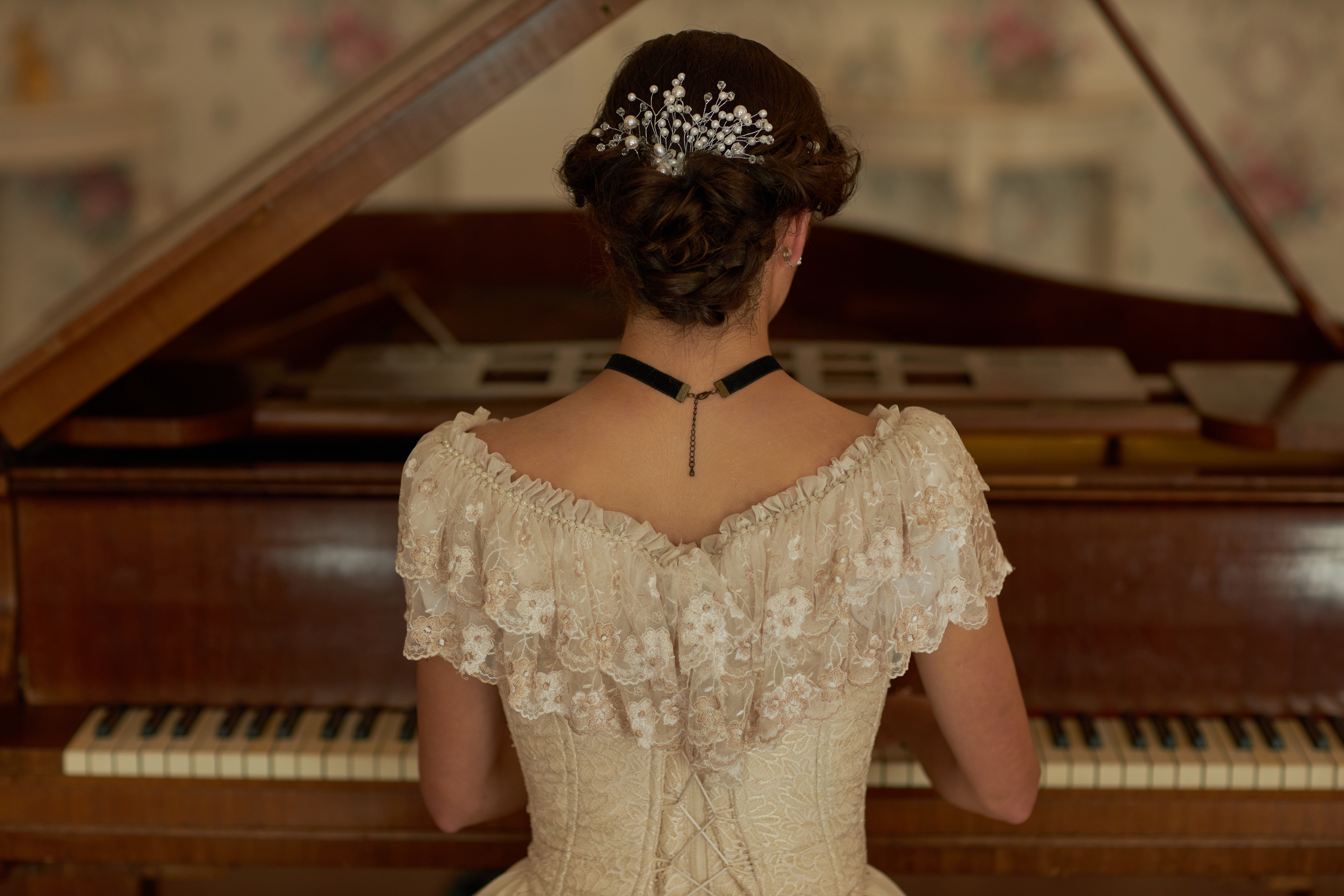 Back view of a person in a lace dress playing a piano, hair adorned with pearls, evoking a vintage or classical style