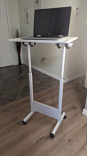 Adjustable standing desk with wheels, holding an open laptop, positioned in a modern room with a tiled and wooden floor
