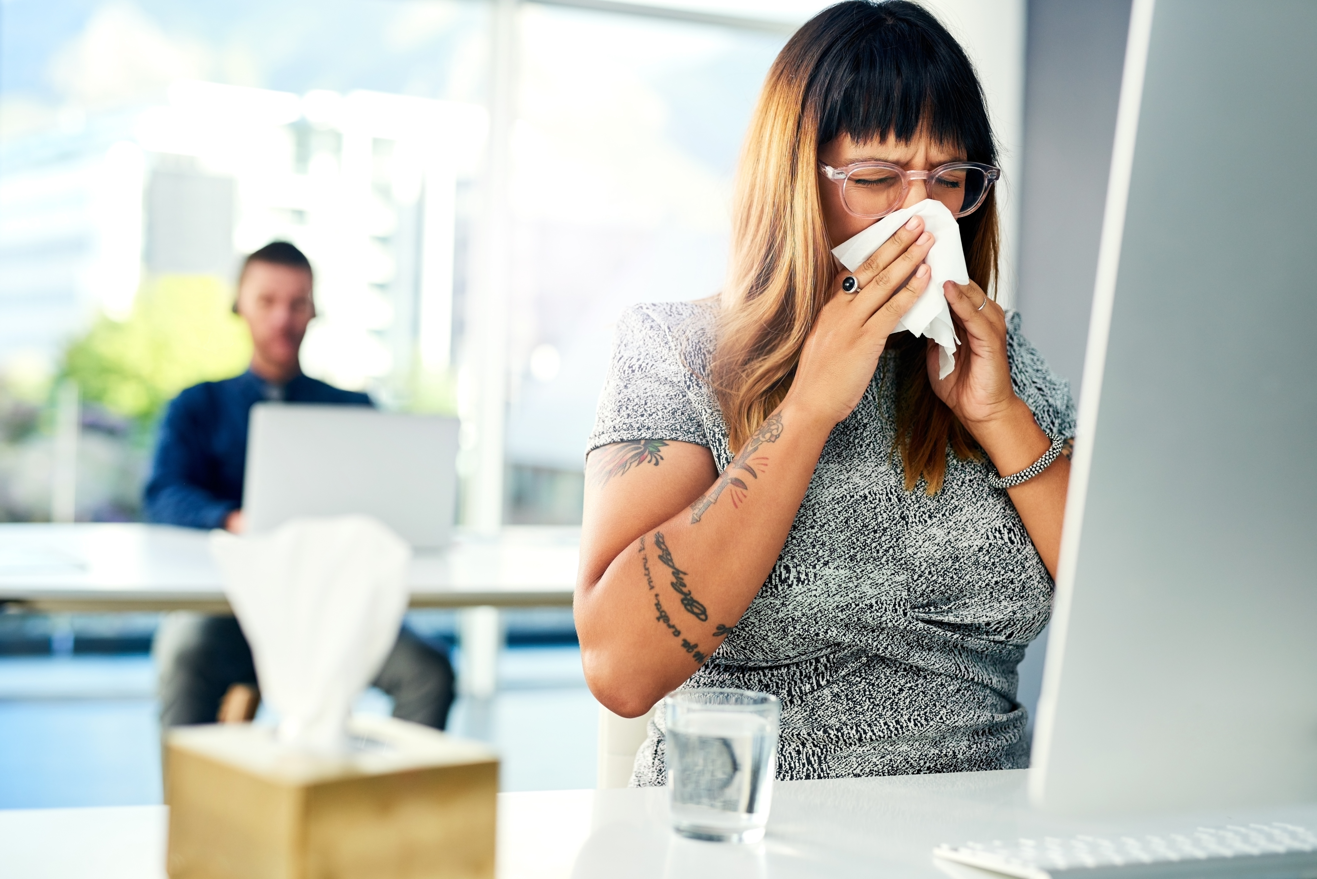 Person with glasses and tattoos sneezes into a tissue at a work desk; blurred colleague in background.