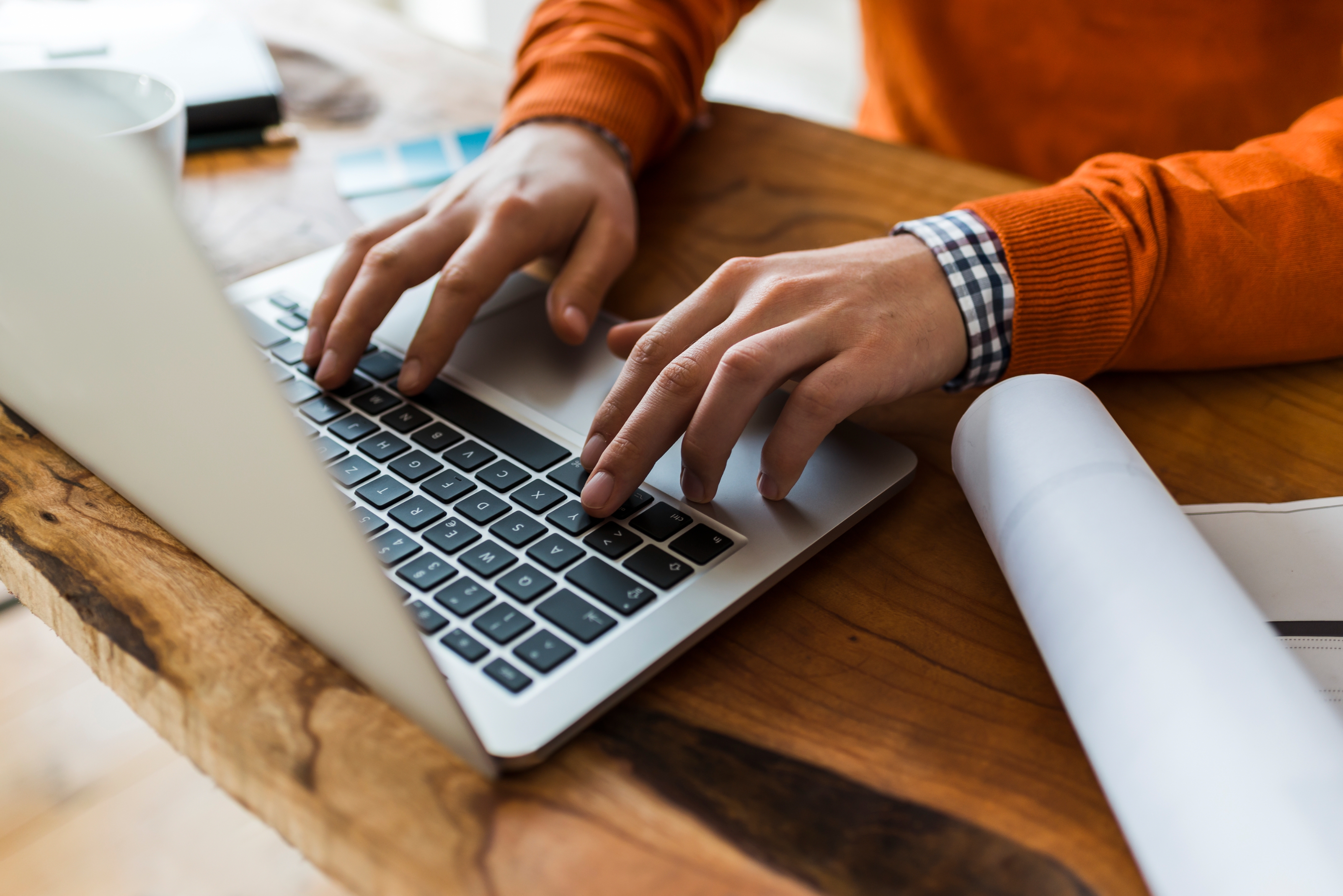Person typing on a laptop at a wooden desk with a roll of paper nearby.