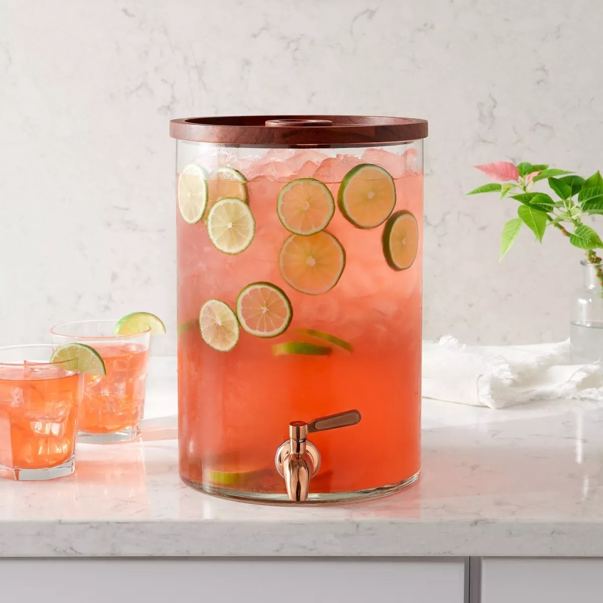 Glass drink dispenser with a spout, filled with iced pink lemonade and sliced limes. Two glasses of the drink are beside it on a marble counter