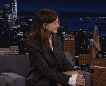 Natalia Dyer on a talk show set, wearing a tailored jacket and sitting in conversation. Cityscape backdrop visible