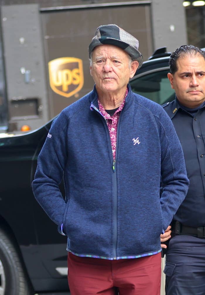 Bill Murray wears a patchwork cap, a patterned shirt, and a blue jacket, walking with a security guard on a city street