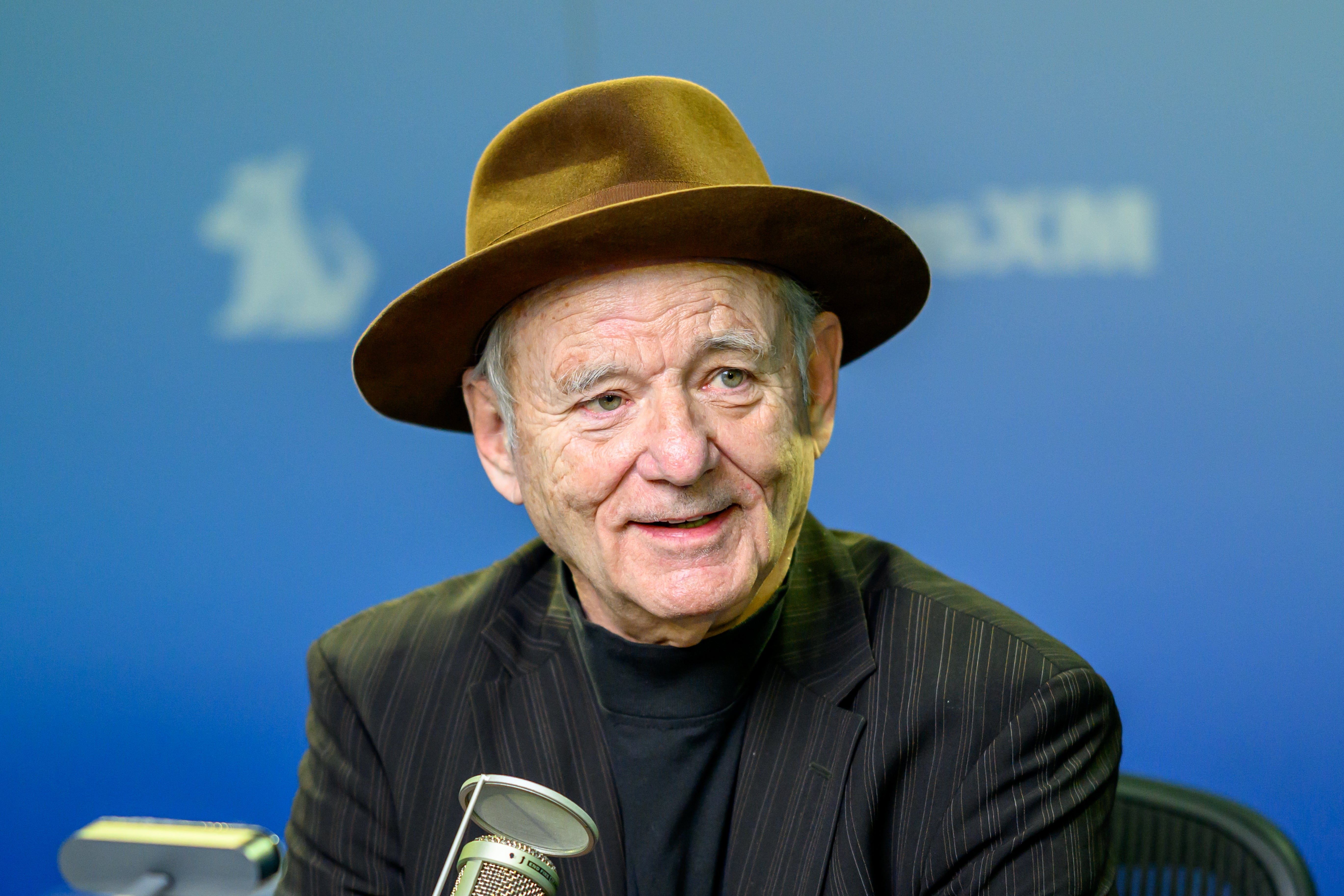 Bill Murray in a pinstripe suit and wide-brimmed hat speaking into a microphone in a studio setting