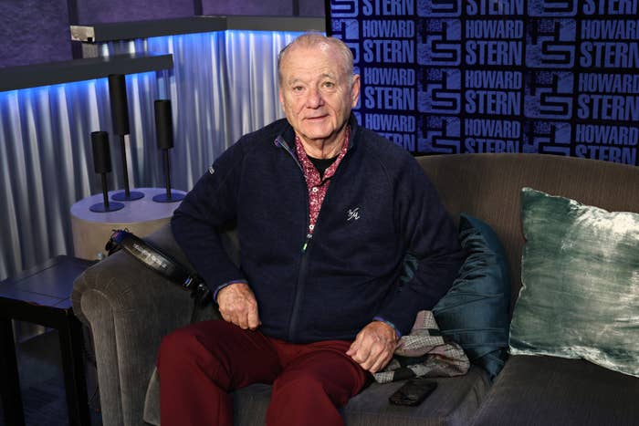 Bill Murray sitting on a couch in a radio studio, wearing a casual zip-up jacket and pants, with a Howard Stern show background