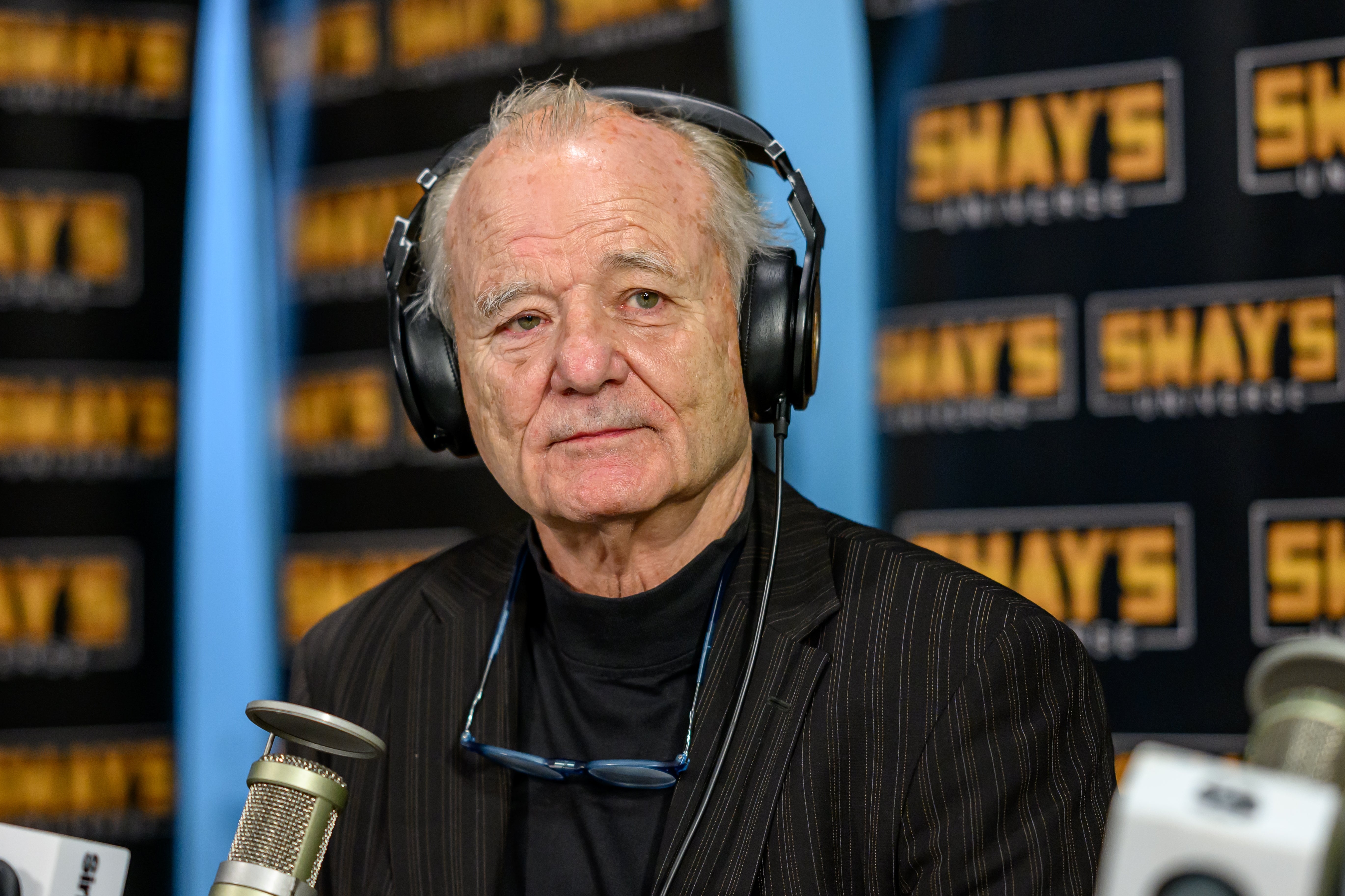 Bill Murray wearing headphones and a striped blazer speaks into a microphone during a radio interview