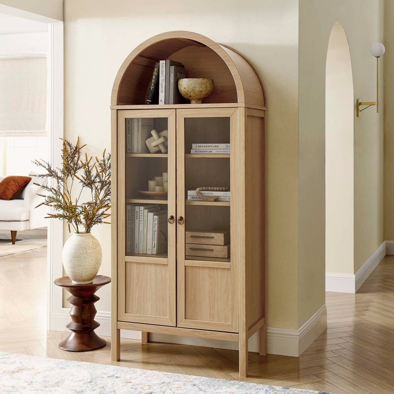 Wooden cabinet with books and decor items, glass doors, beside a small table with a vase. Bright, elegant home interior setting