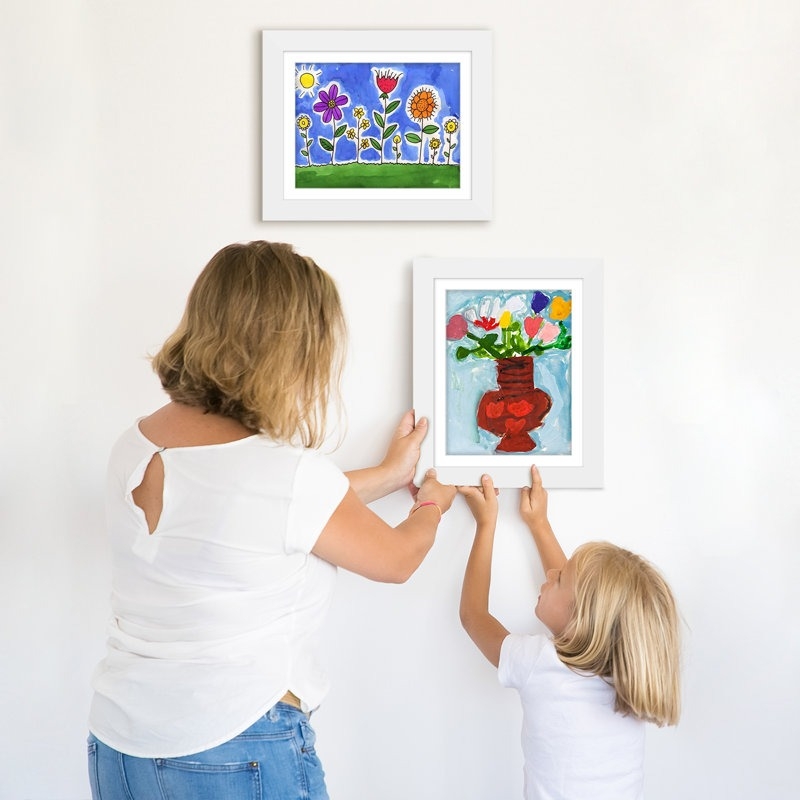 Adult and child hang colorful, framed floral art on a white wall, showcasing creative home decor options