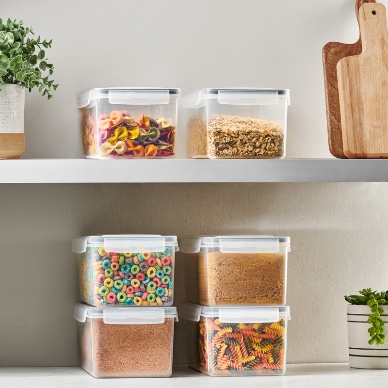 Clear plastic storage containers on a kitchen shelf, filled with various dry foods like cereal, pasta, and grains. A potted plant and cutting boards are nearby