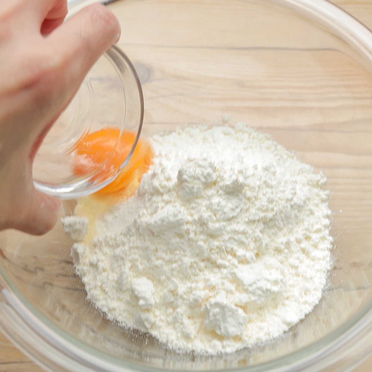 手がボウルに卵黄を小さなガラス容器から小麦粉に注ぎ入れている。調理の準備工程の一部。