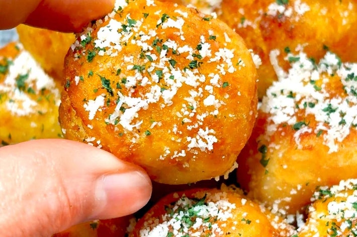 手で持たれたチーズとハーブがかかった焼きポテトボールのクローズアップ。