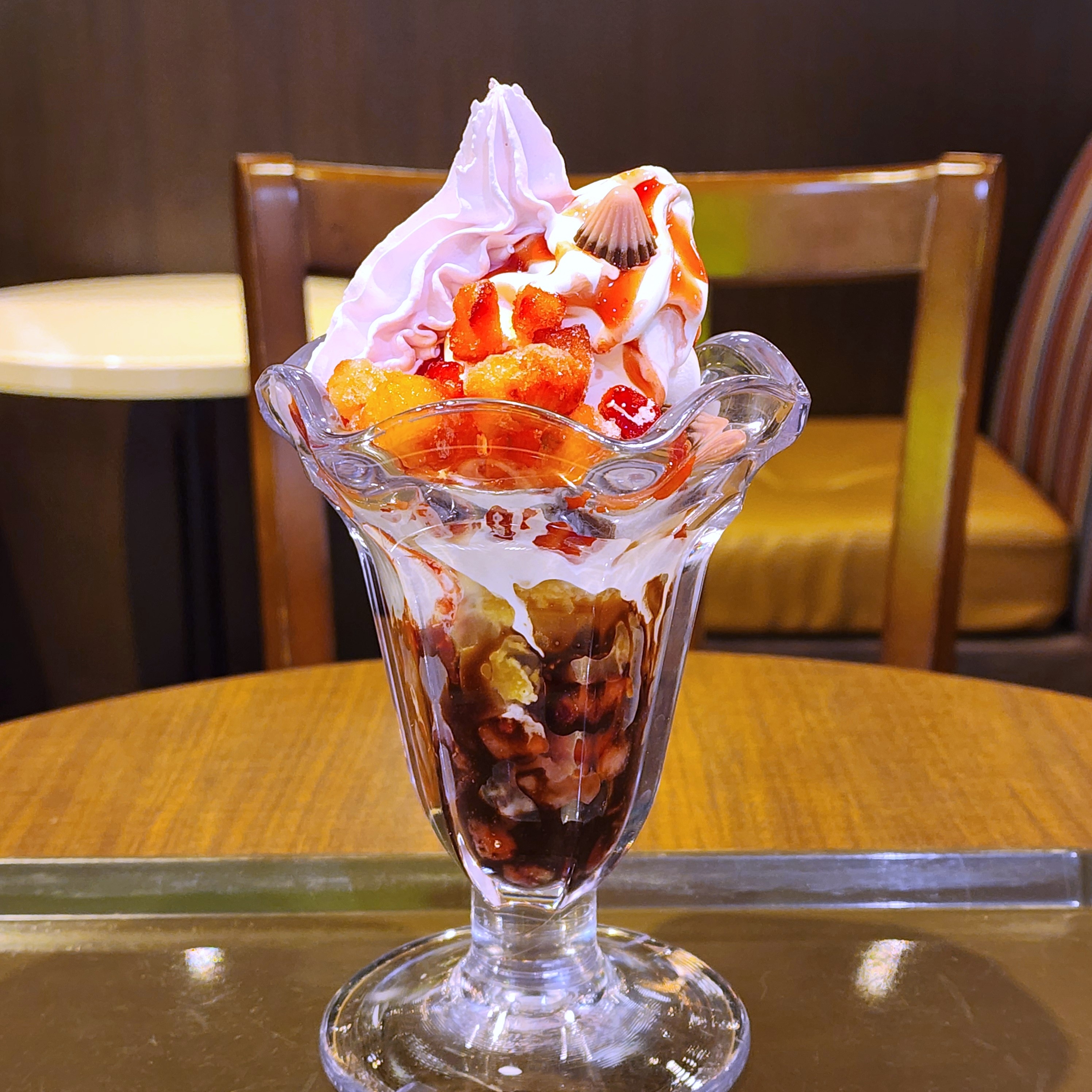 パフェがテーブルに置かれている。クリーム、フルーツ、チョコレートソースがトッピングされ、美味しそうに盛り付けられている。