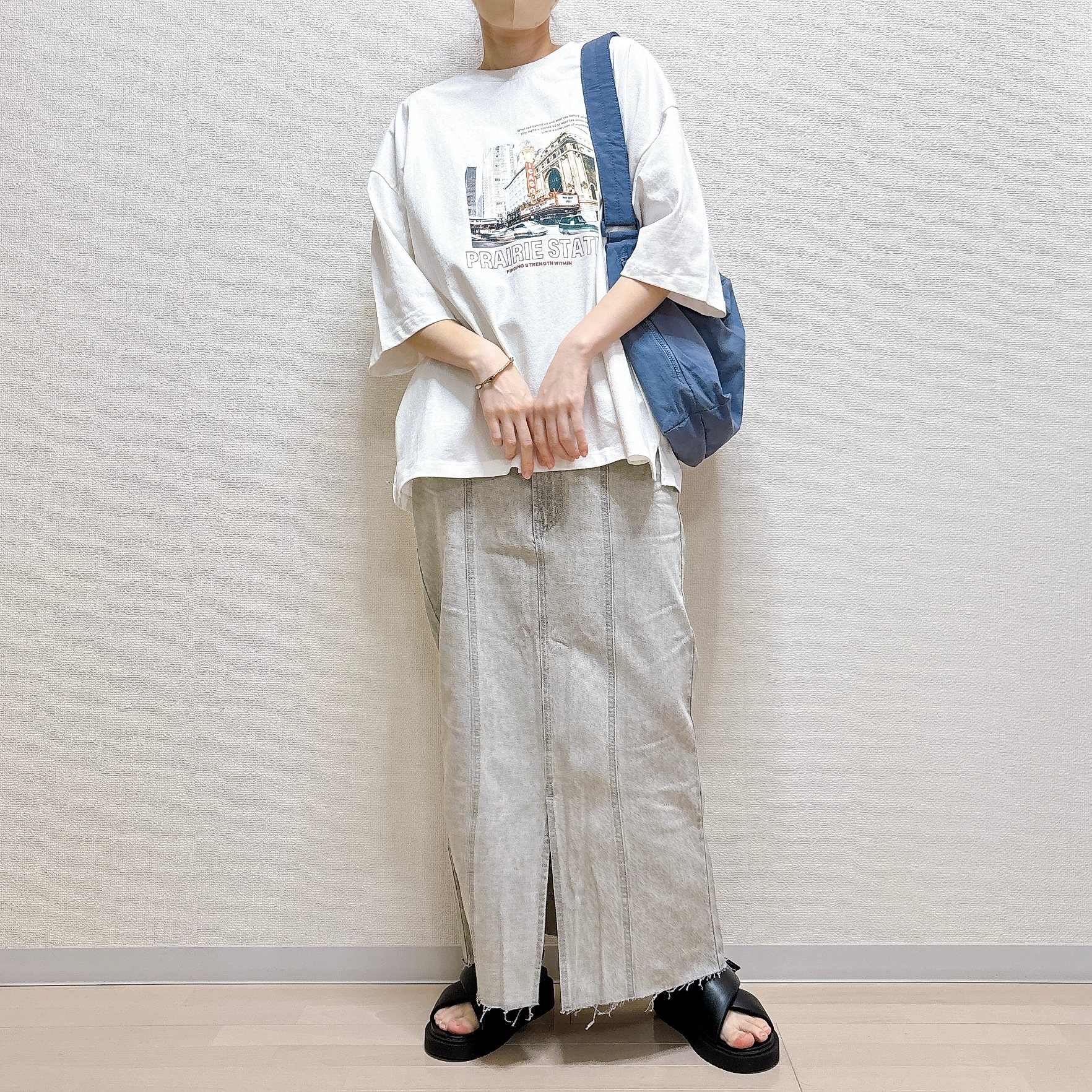 ゆったりとしたTシャツとロングスカートを着た人が、バッグを肩に掛け、カジュアルなサンダルを履いて立っている。