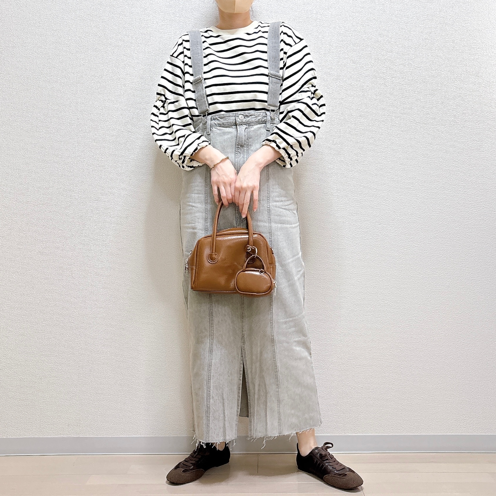 人物がストライプのシャツとサロペットを着て、茶色のバッグを持っている。靴はカジュアルなスタイル。