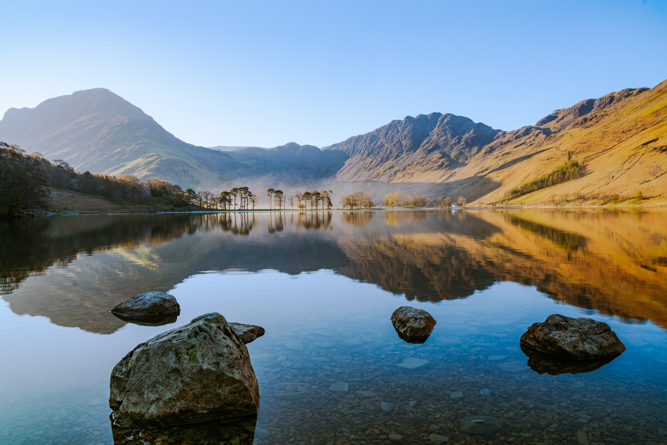Serene lake scene with mountains reflecting on calm water, large rocks in the foreground, and a line of trees in the distance