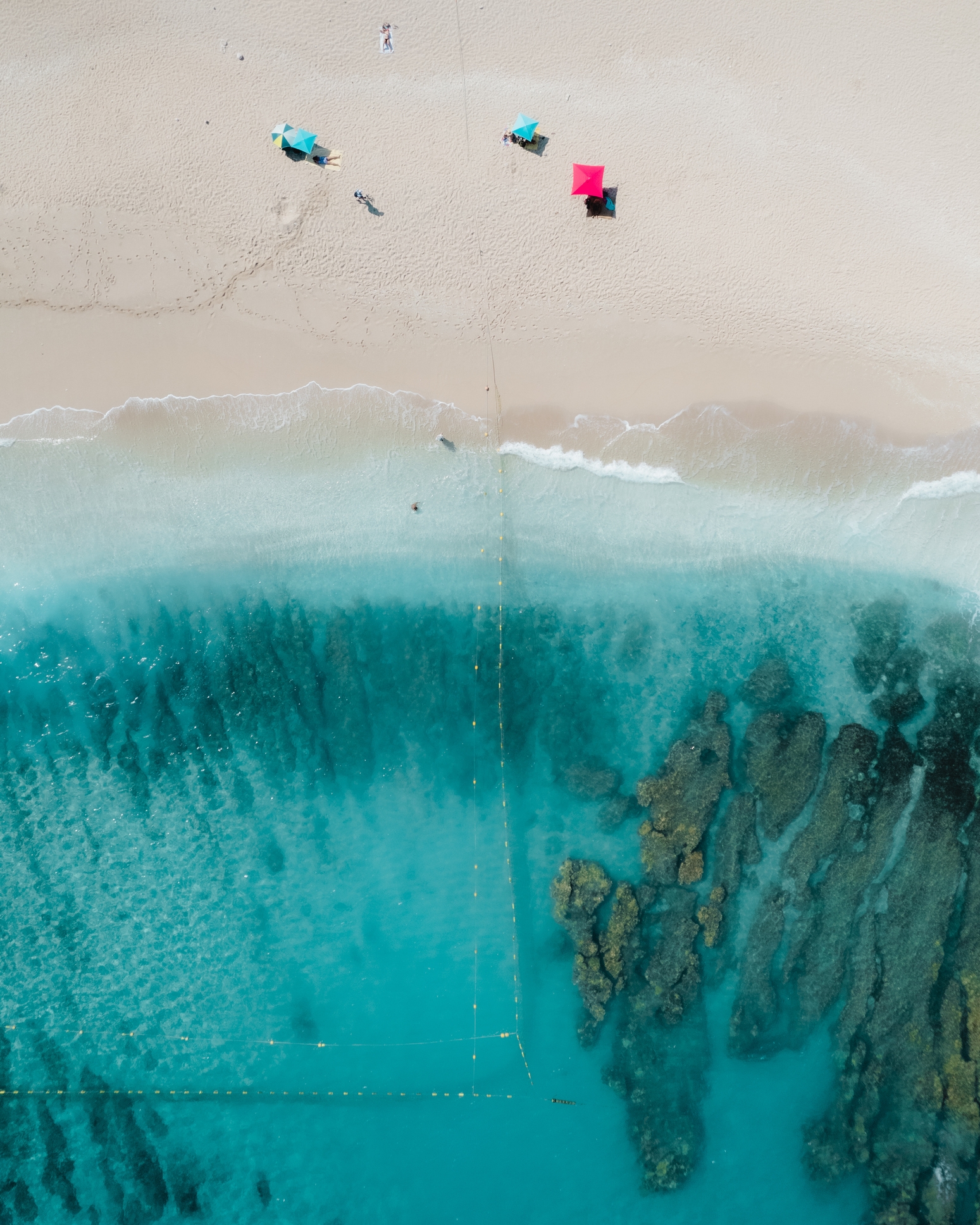Aerial view of a beach with people under umbrellas by the shore and clear water, revealing underwater rock formations