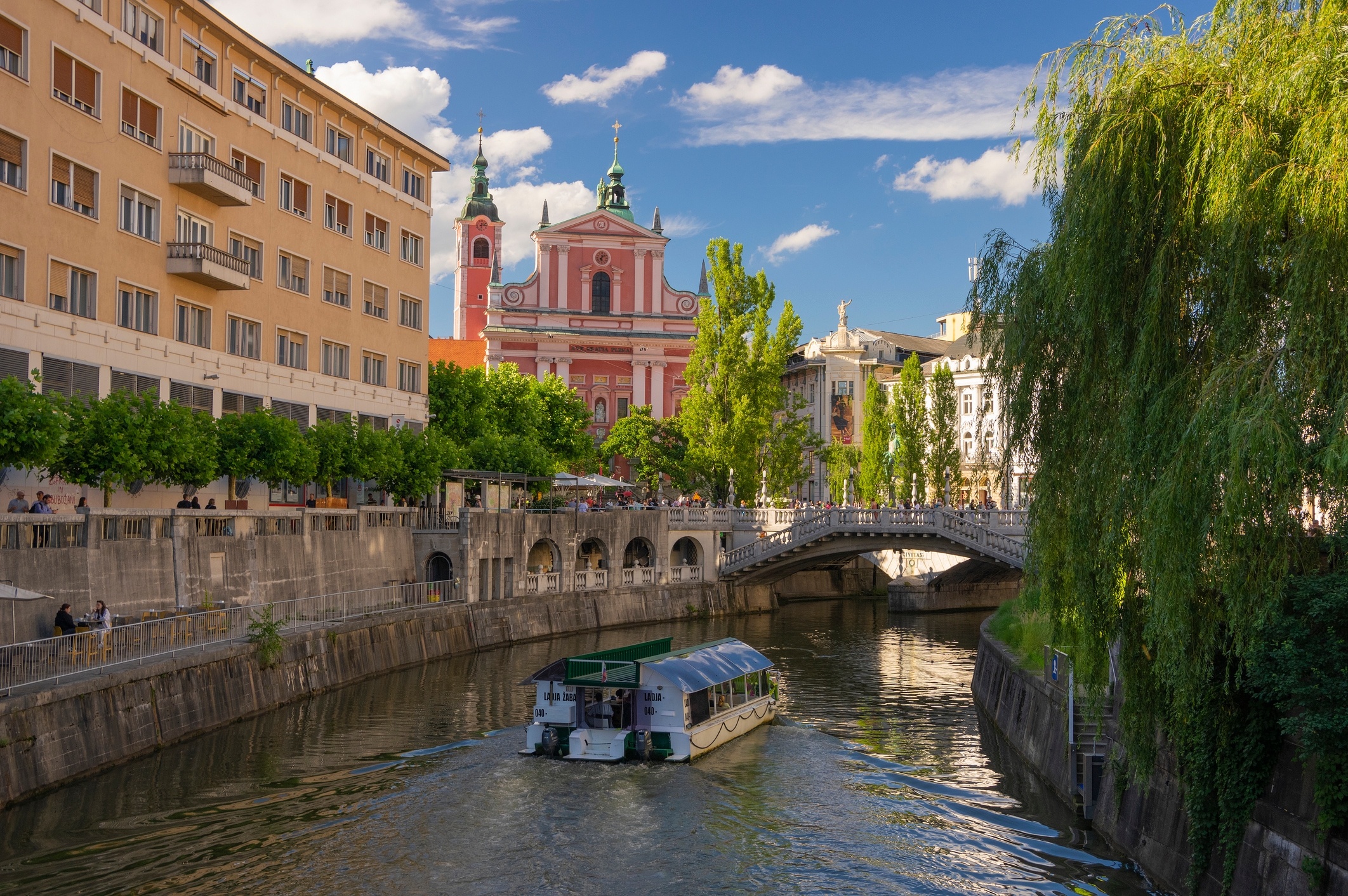 A scenic canal in a European city, framed by historic buildings and a bridge in the background; a boat cruises along the tranquil water