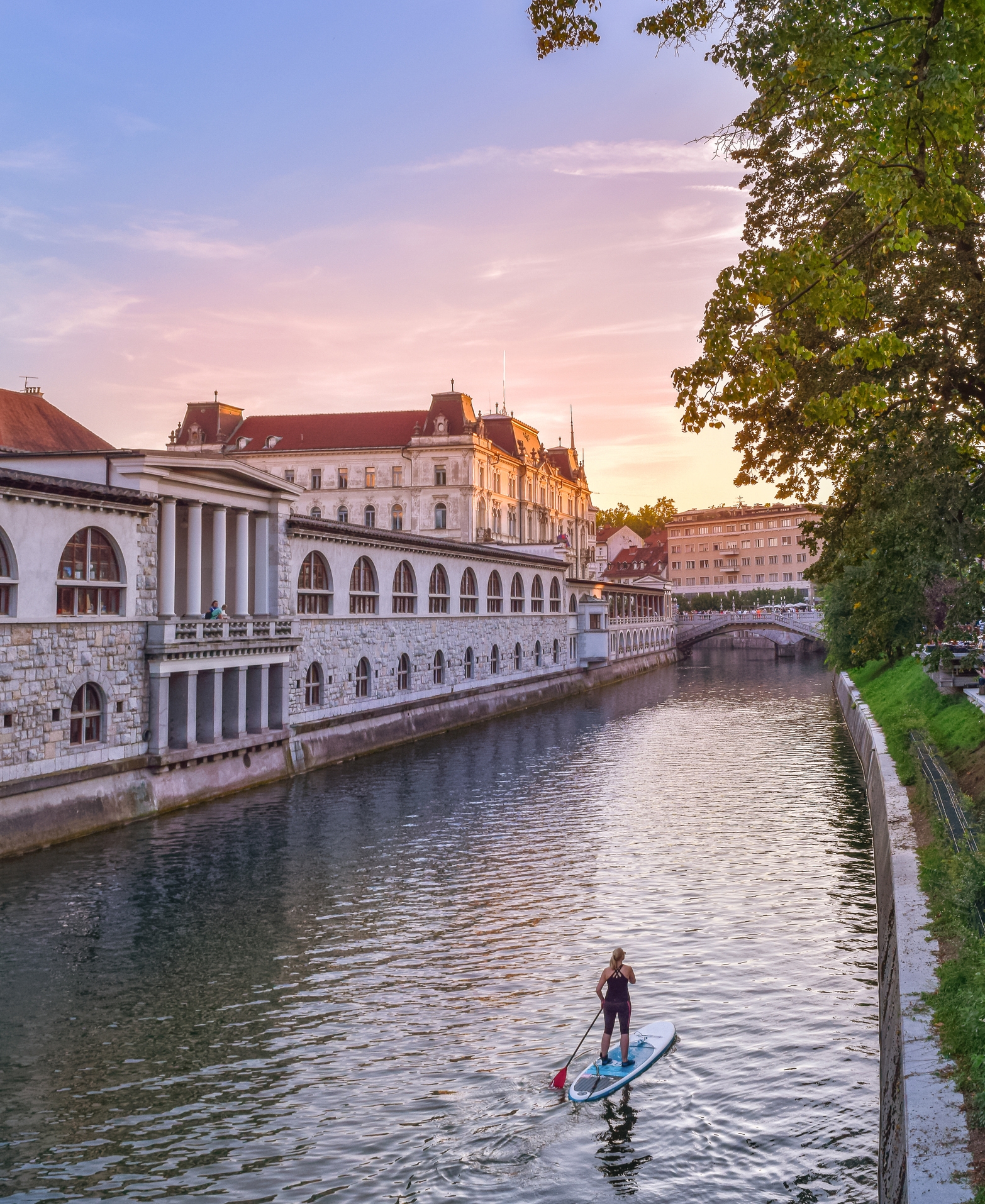 Person paddleboarding on a river through a European city, with historic buildings and trees lining the riverbank at sunset