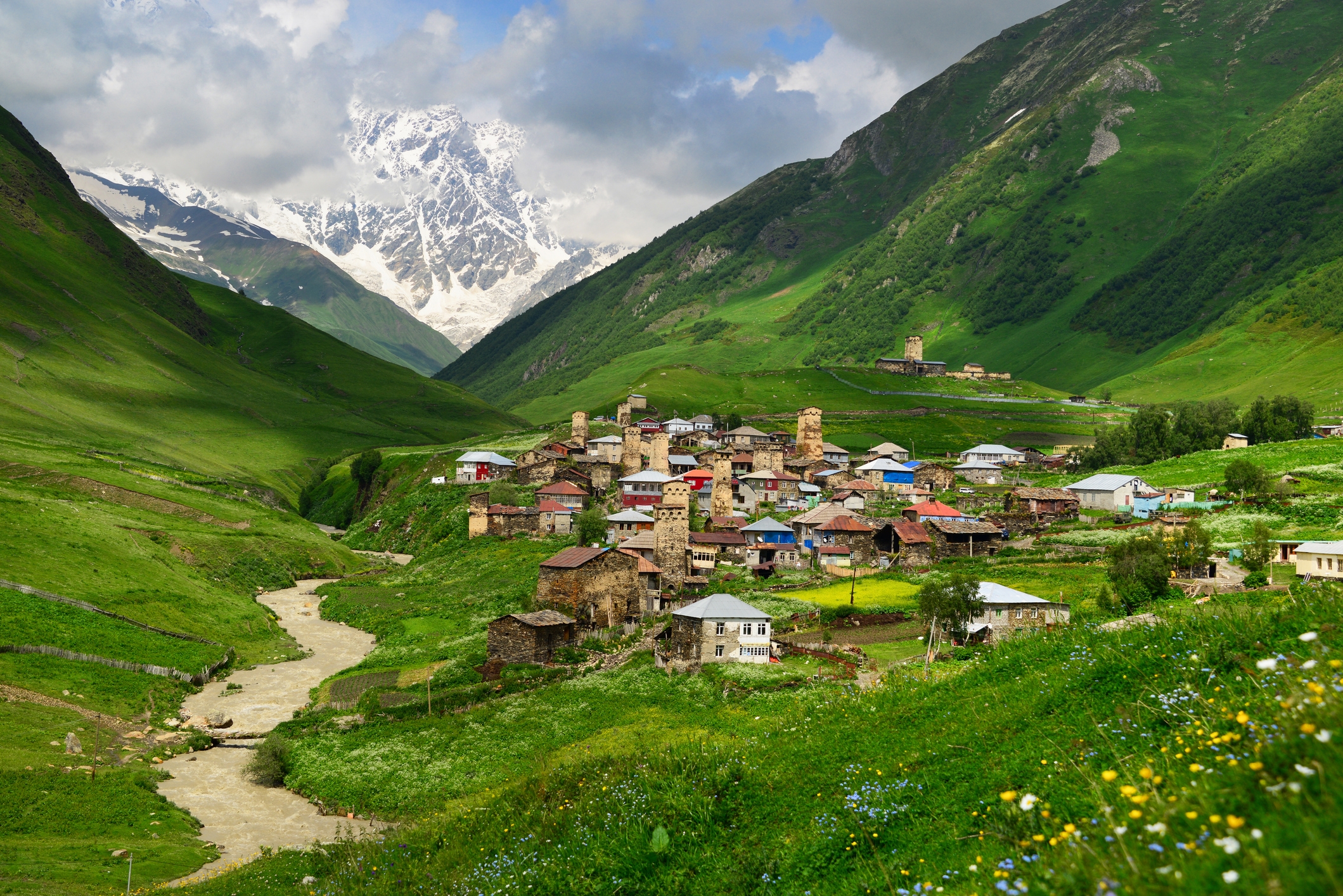 Mountain village with stone houses nestled in a lush green valley, surrounded by towering mountains and a winding river