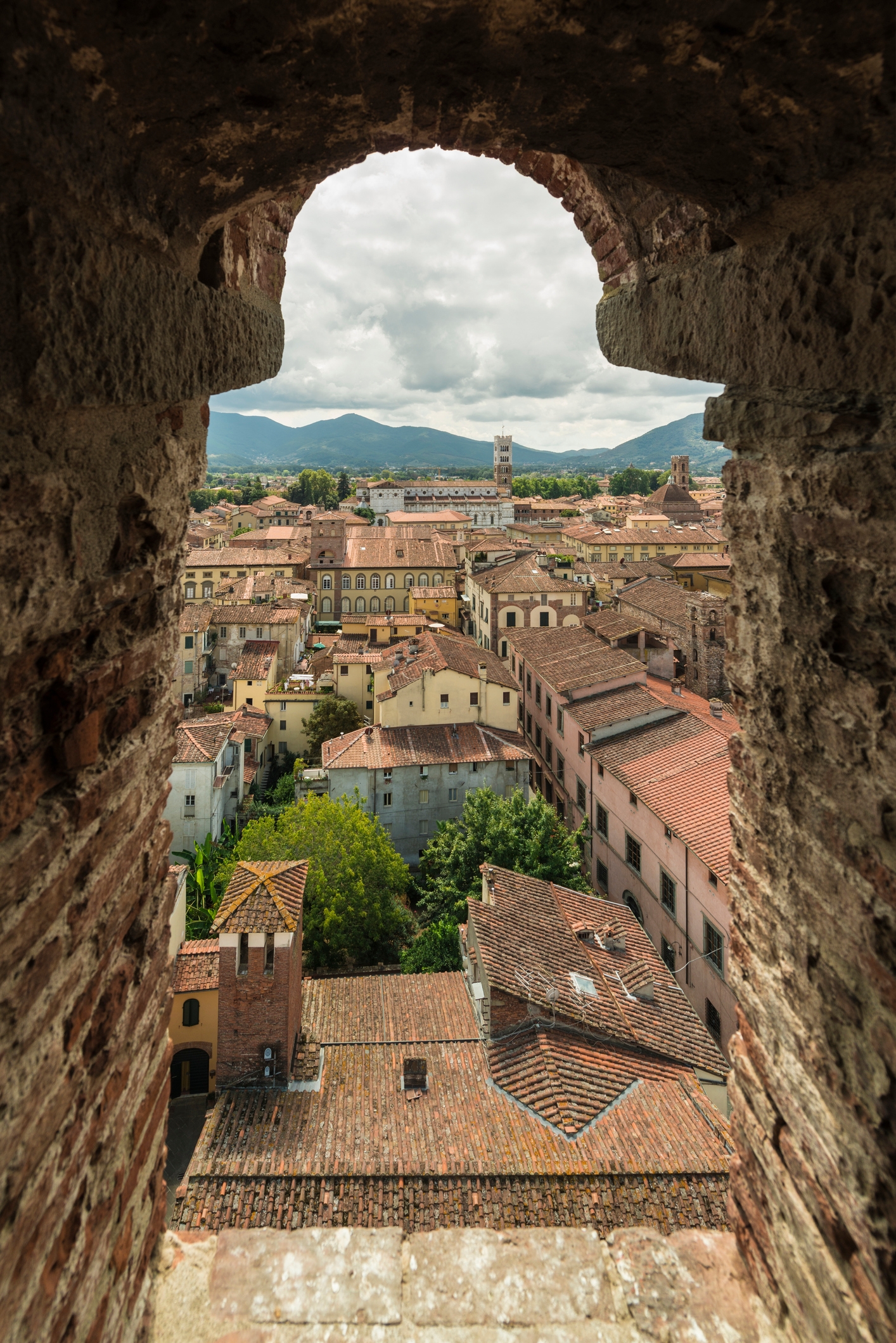 View through a stone archway overlooking a historic town with terracotta rooftops and distant rolling hills under a cloudy sky