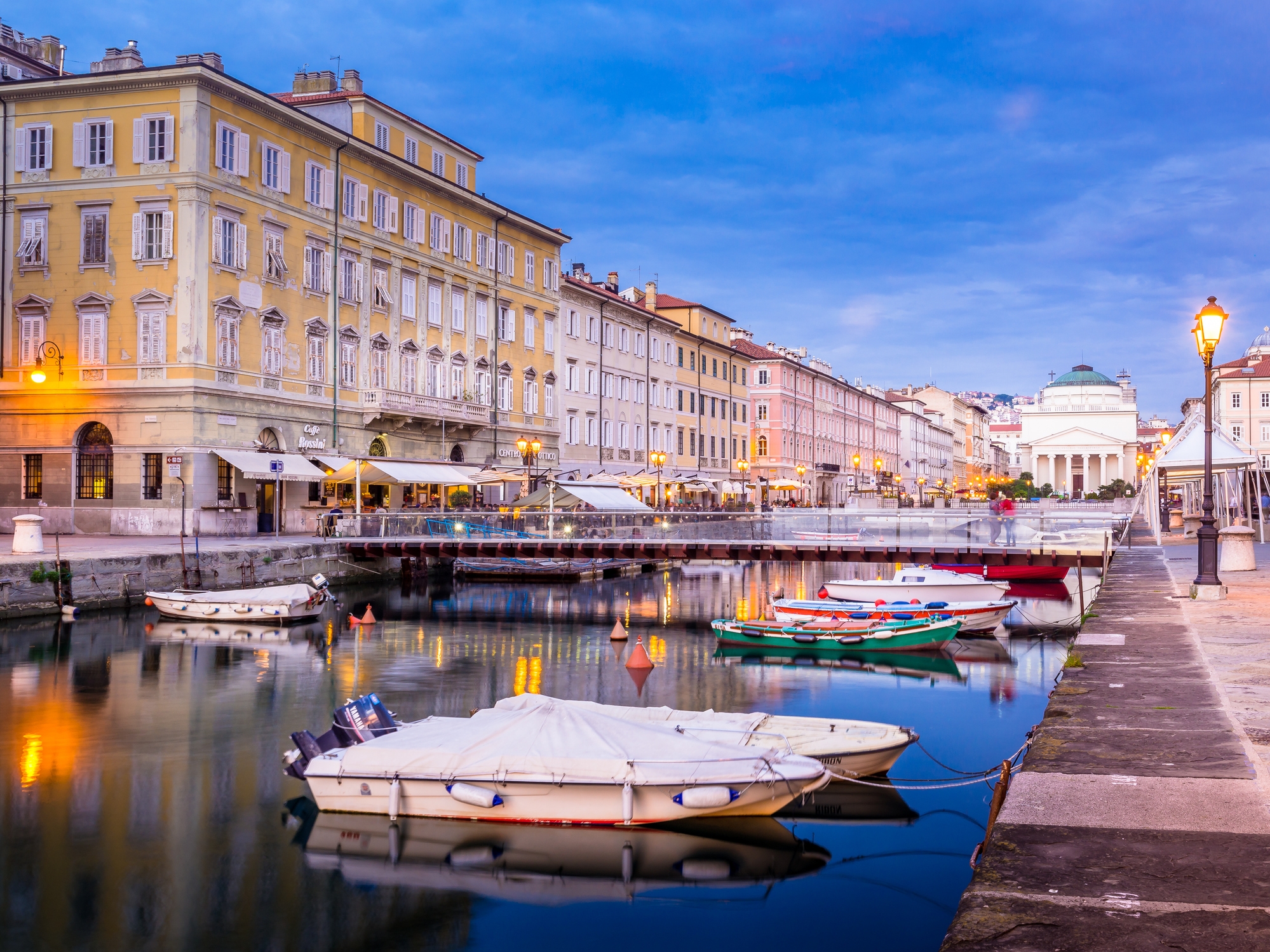 Canal lined with moored boats and historic European architecture, leading to a domed building in the distance under a twilight sky