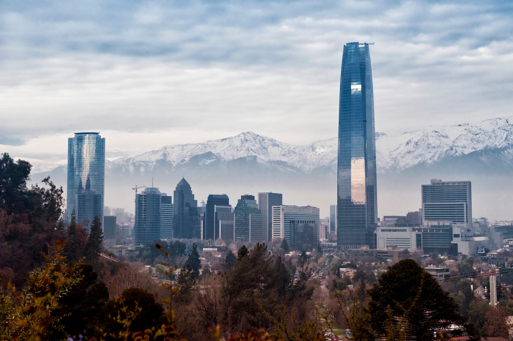 A city skyline with modern skyscrapers, including a tall, reflective building, set against snow-capped mountains under a cloudy sky