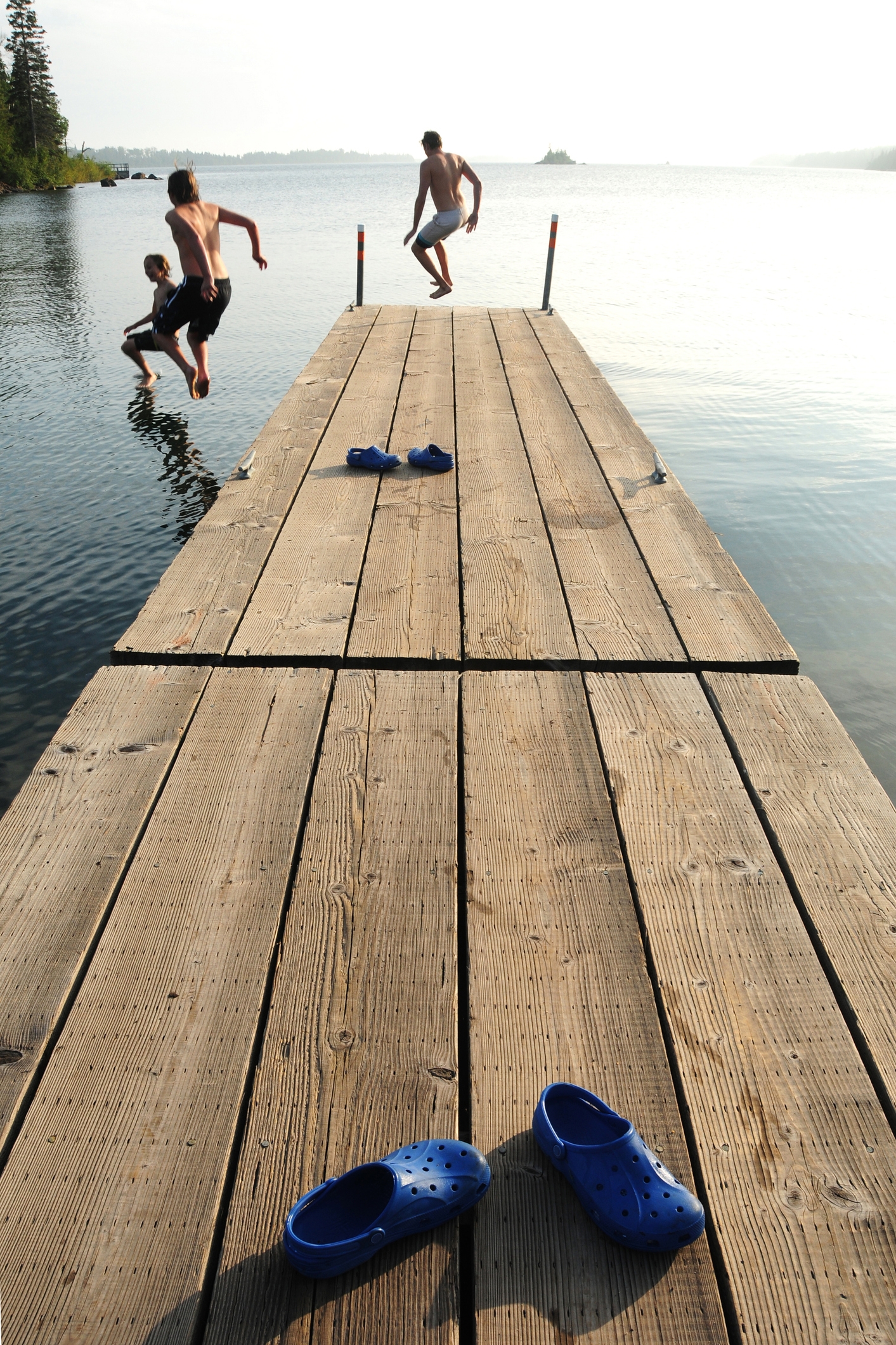 Three people jump off a wooden dock into a lake on a sunny day. Blue shoes are left behind on the dock