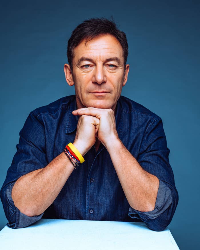 Person in denim shirt rests chin on hands, wearing multiple bracelets on left wrist, against a plain background