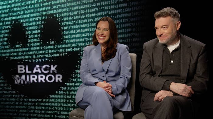Two people are seated for an interview with "Black Mirror" visible in the background. The woman wears a tailored suit, and the man is in a sweater and shirt