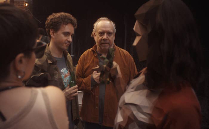 Group of people talking casually; one man holds drink, another wears open jacket, and a few others engage in conversation nearby
