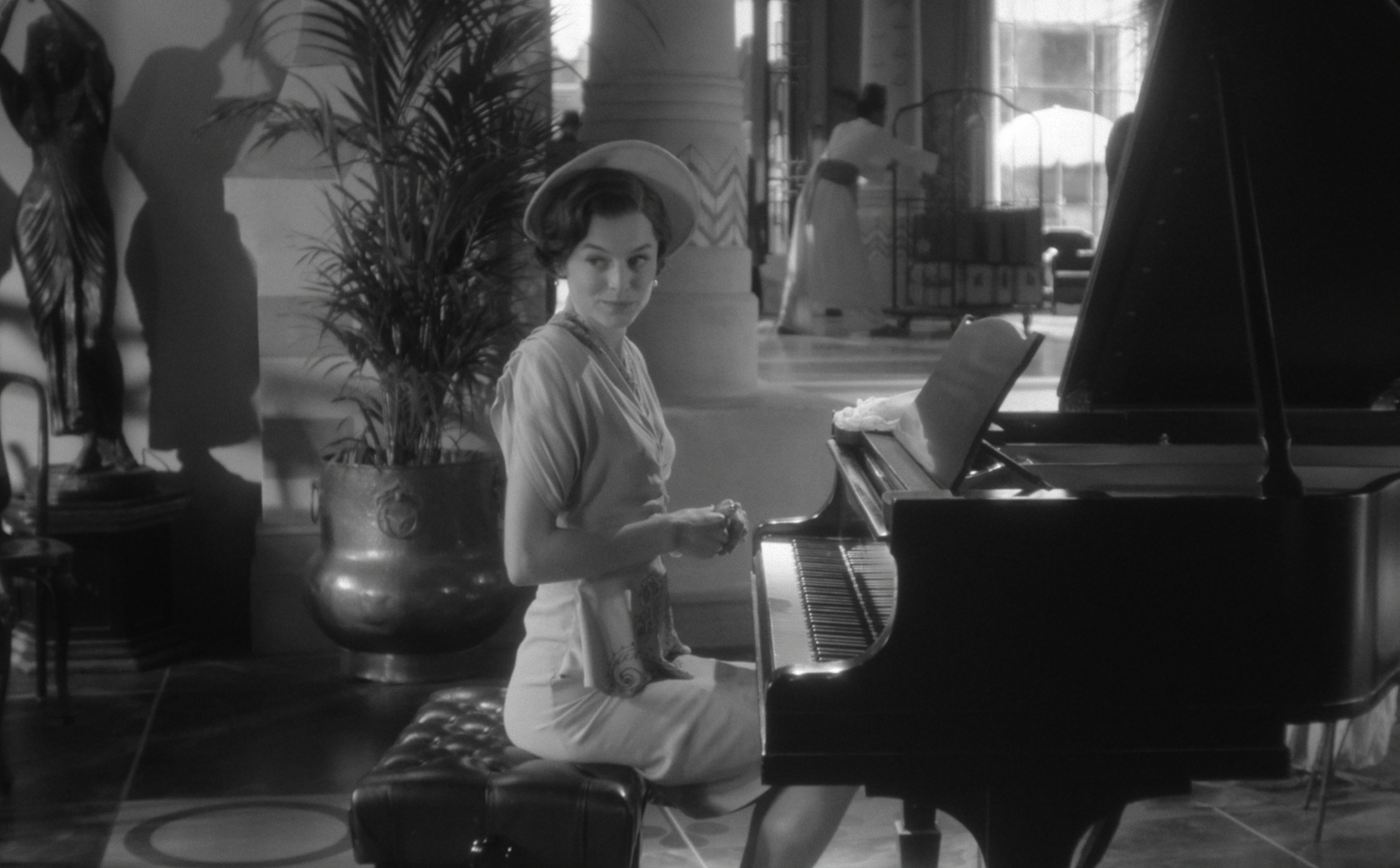 A woman in elegant attire sits at a grand piano in a spacious room, glancing back with a soft expression. Artful decor is visible in the background
