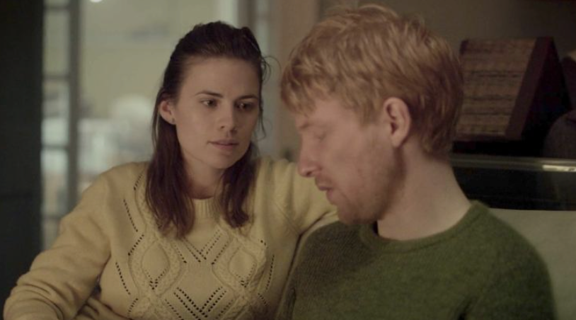 A woman in a knitted sweater looks at a man in a crewneck sweater, both appear reflective and serious indoors