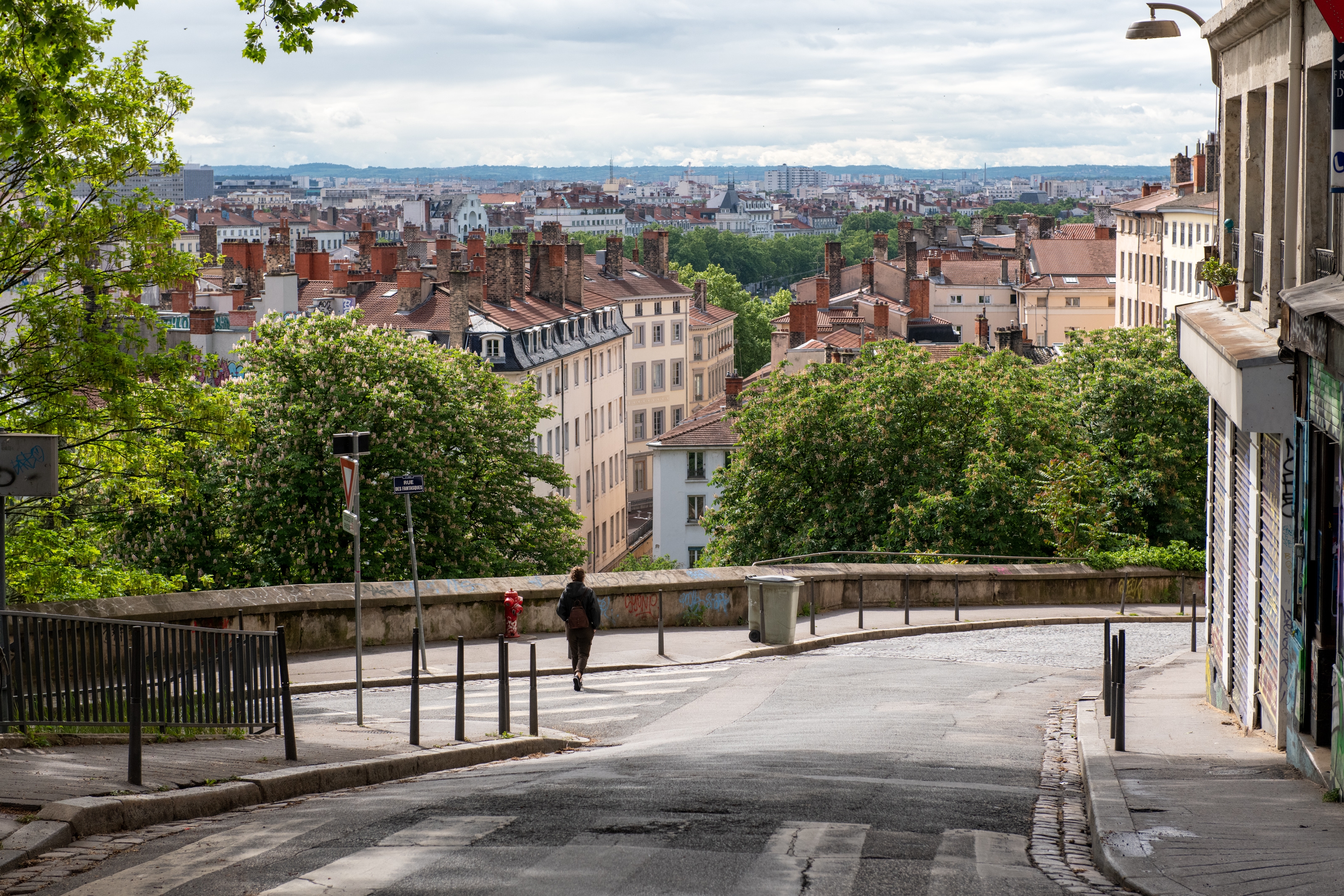 A lone person walks down a quiet street with a scenic view of a cityscape and lush green trees