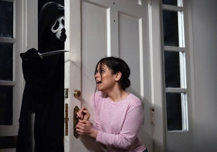 A person in a hoodie and mask menaces Jenna Ortega behind a door inside a house