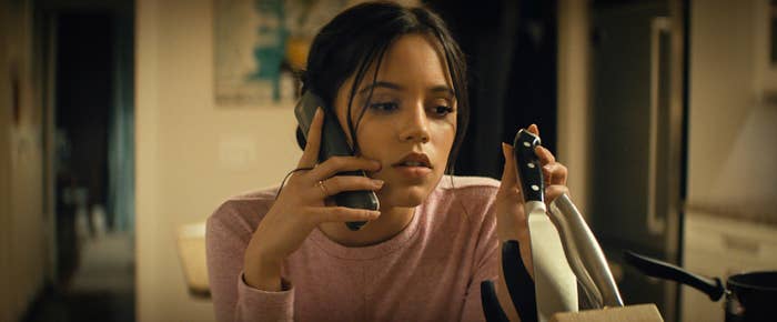 Jenna Ortega in a casual setting holds a phone to their ear, appearing focused, while holding a large knife in the other hand