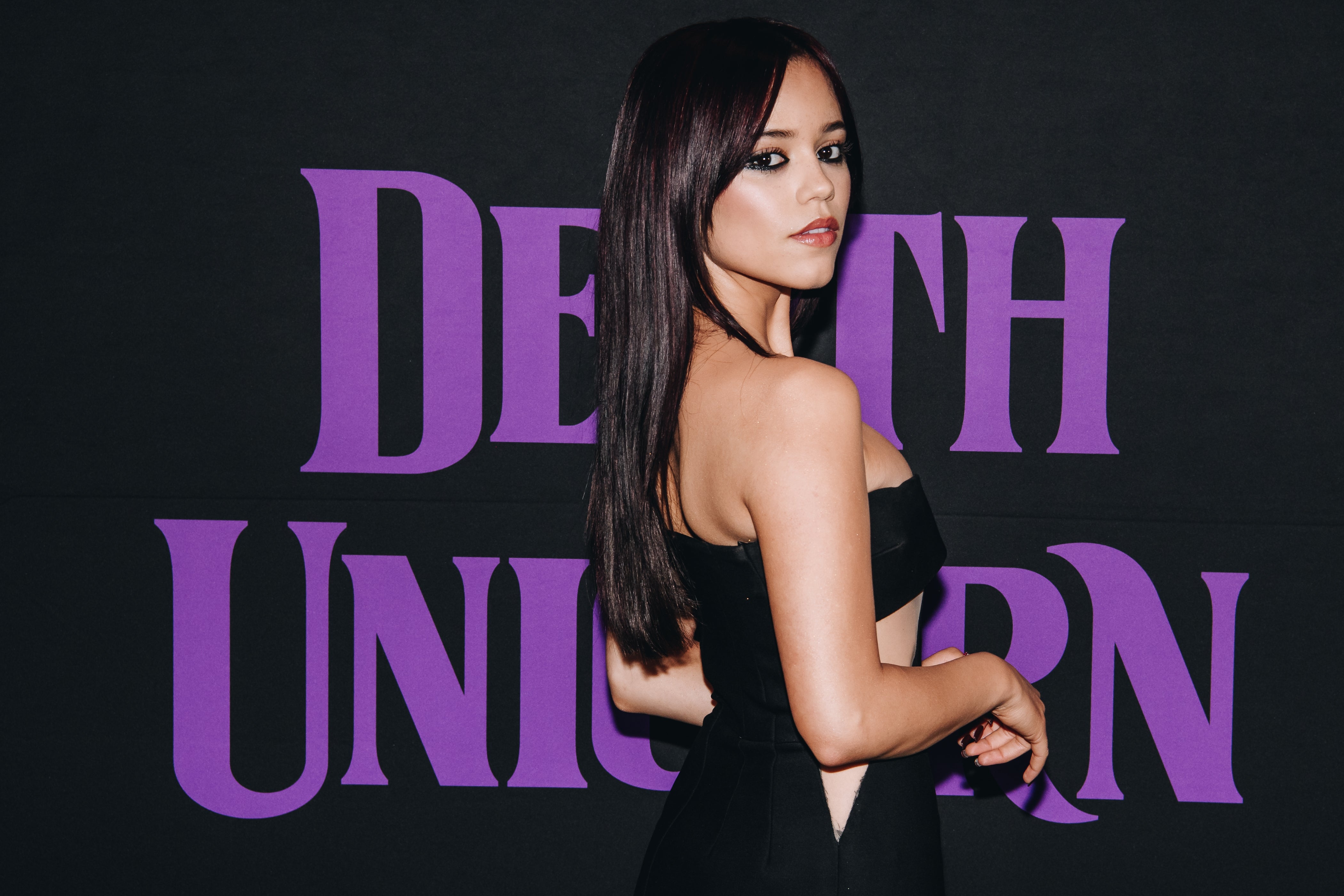 Jenna Ortega in elegant strapless dress poses in front of a backdrop with text "DEATH UNICORN."