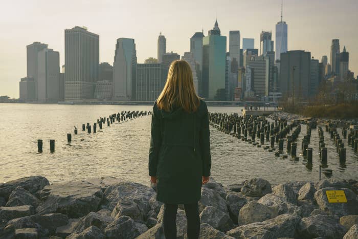Person stands on rocky shore, facing city skyline with skyscrapers across the water