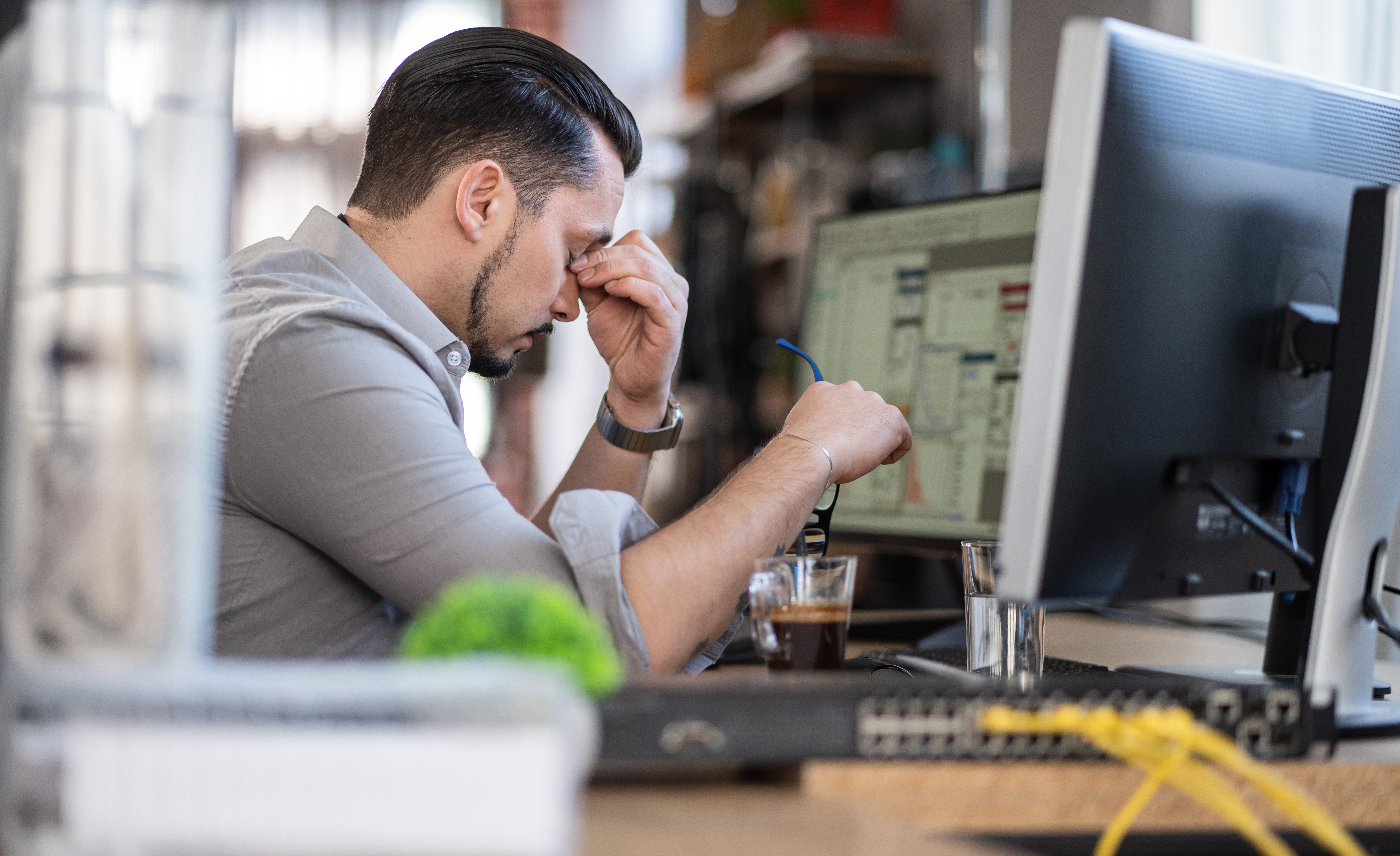 A person sits at a desk, looking stressed, holding glasses, in front of a computer screen with blurred content